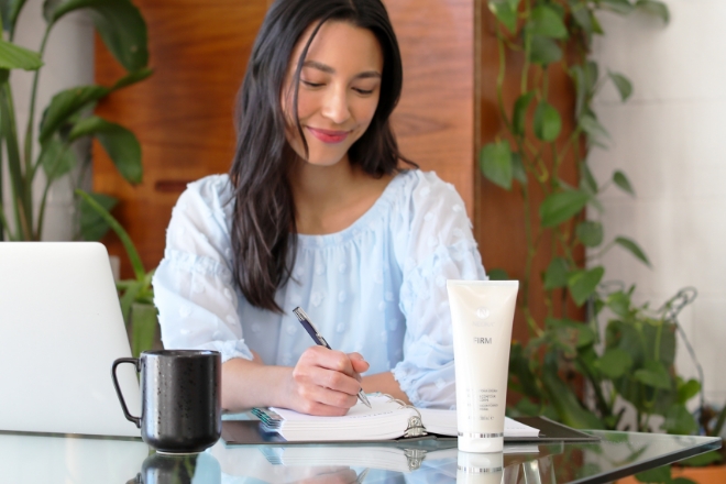 Image of woman working with laptop, writing In a notebook with a bottle of Firm Body Contour Cream on the table.
